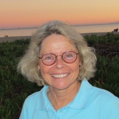 Kathleen Komar wears a teal polo shirt and stands on a beach with the sunset in the backgrounf.