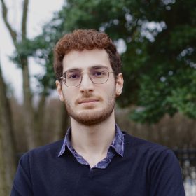 Headshot of Askaan Kashani wearing a navy sweater and standing in front of green foliage.