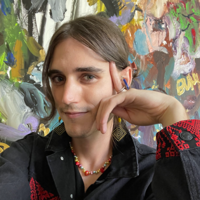 Isaac Dwyer wears a navy jacket with red embroidery, seated in front of a colorful abstract painting.