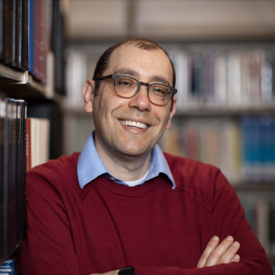 Headshot of Gideon Manning in a red sweater, leaning on a bookshelf.